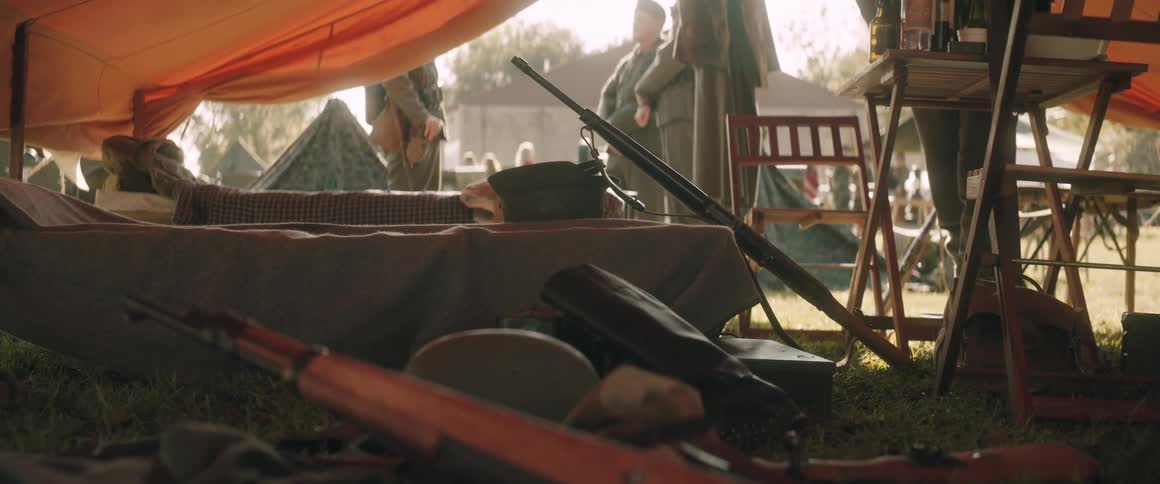 Military tent with bed rifle and gear on the ground with soldiers and structures in the background during daytime