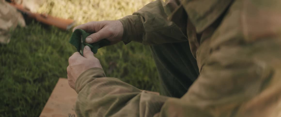 Person in brown shirt cleaning item beside box and rifle on grass