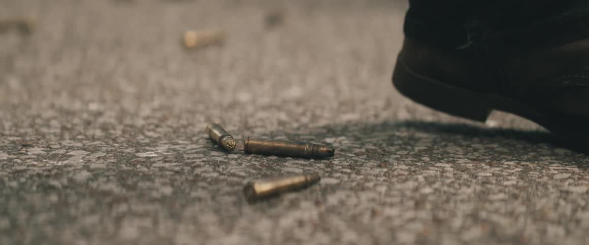 A closeup shot of spent rifle bullet casings scattered on a textured grey ground with a persons boot briefly stepping into the frame near the casings