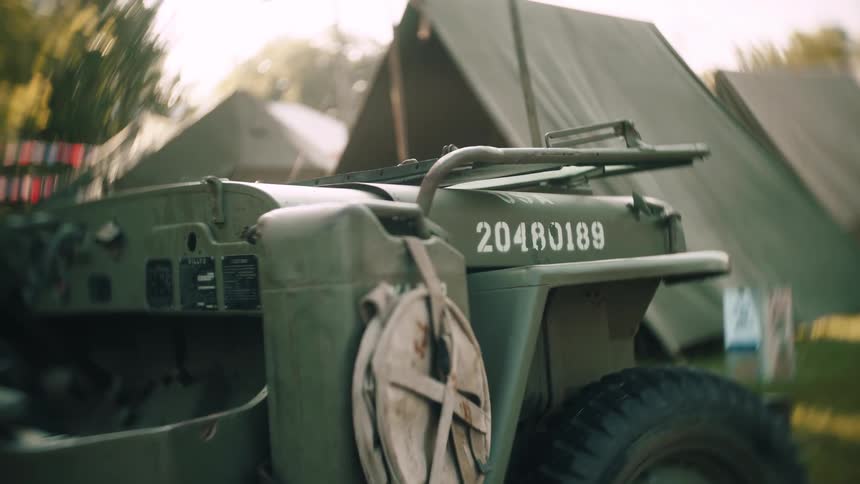 A vintage olive drab military jeep is parked amongst large canvas military tents at a sunny outdoor camp