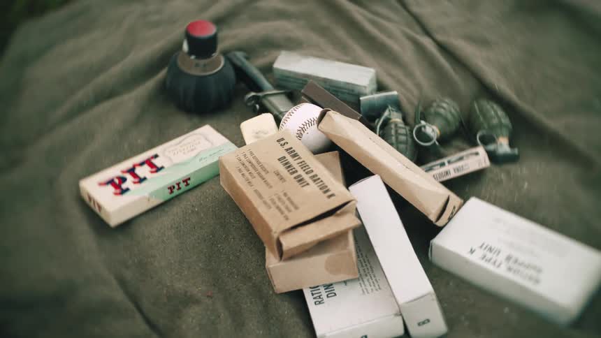 Various military gear and personal supplies including tree piece of grenade and a baseball arranged on a service members bed at midday