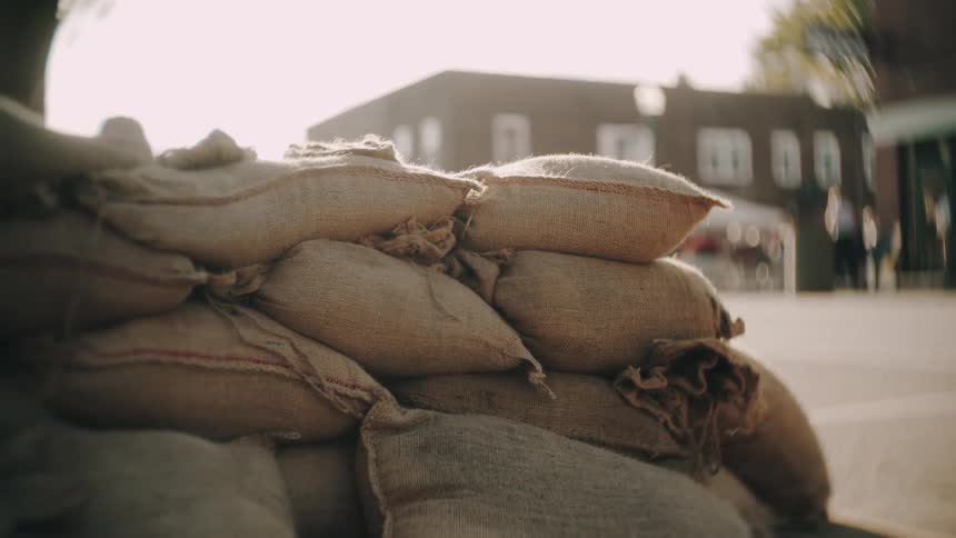 A defensive barricade constructed from stacked burlap sandbags heavily backlit by bright sunlight with an outoffocus background showing blurred dark brick buildings and distant figures in an urban setting