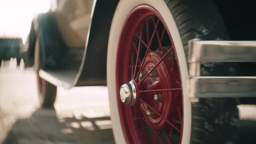 The vibrant red spokes and whitewall tire of a vintage car spin rapidly in a detailed closeup the chrome hubcap glinting in the sunlight as the vehicle travels down the road