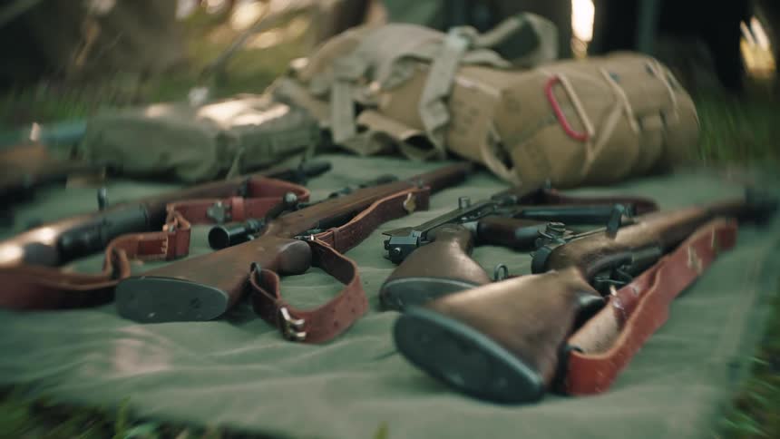 An assortment of vintage rifles and military equipment rests on a green canvas tarp on the grass in a forest camp