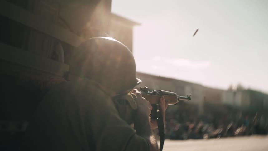 Military soldier stands near a building and fires his gun on a street and shell casings fly into the air