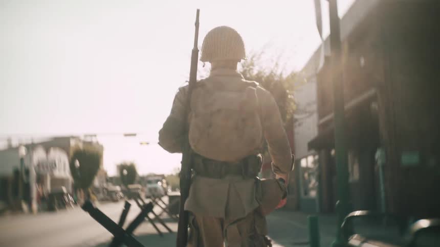 A lone soldier dressed in full combat gear walks away down a desolate town street lined with defensive antitank obstacles his silhouette outlined by the blinding glare of the sun