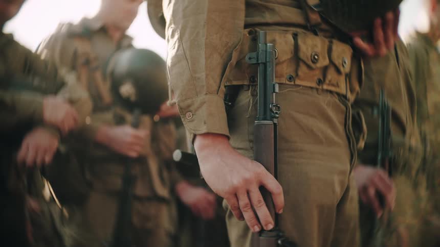A closeup shot focuses on a soldiers hand on his rifle as he stands at attention with his platoon their vintage uniforms visible in the sunlight