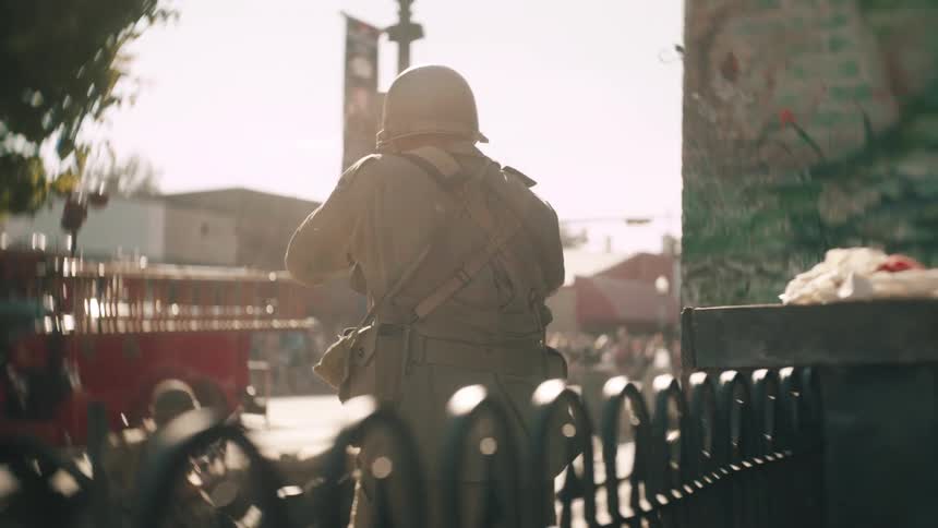 A soldier in an old fashioned uniform and helmet seen from behind walks aims and fires his weapon on a busy sun drenched street with a red fire truck in the background