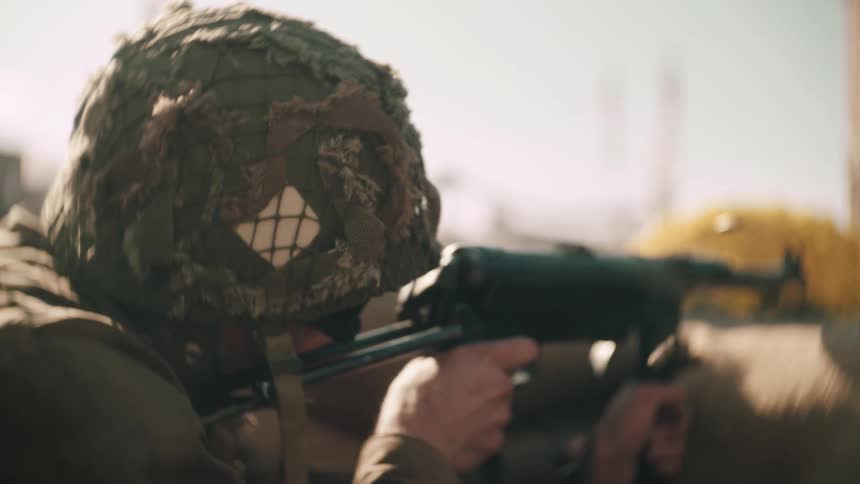 Military soldier stands outside and fires their gun on a street and shell casings fly into the air