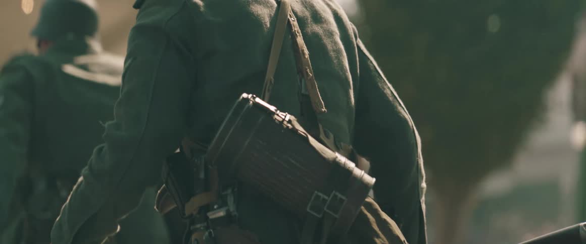 A man in a green military jacket and a gas mask container slung over his shoulder runs across a dusty outdoor area with blurred figures in the background
