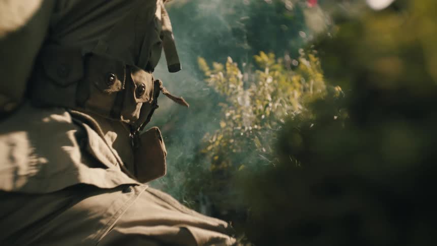 Amidst the chaos of battle a soldier in vintage gear crouches behind a bush and pulls the pin on a smoke grenade and throws it