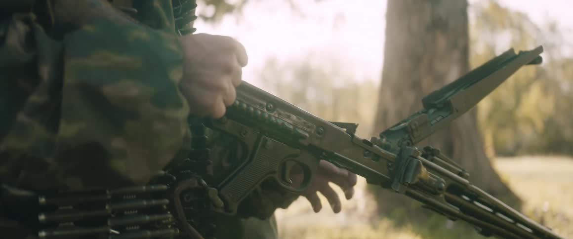 A man in a green camouflage jacket loads an ammunition belt into a machine gun in a sunny forested outdoor area