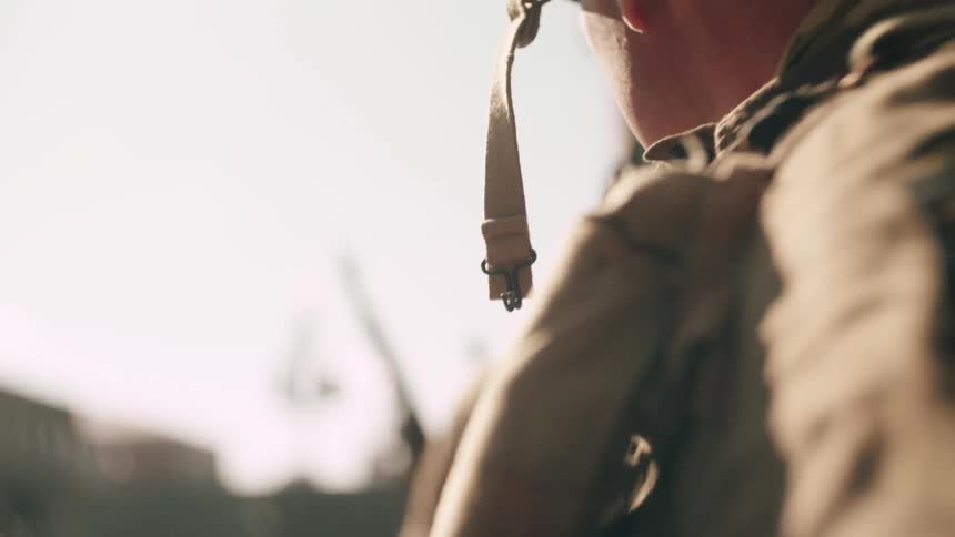 A military officer wearing a helmet and uniform sitting on the ground behind a wall at midday taking cover and observing the surroundings