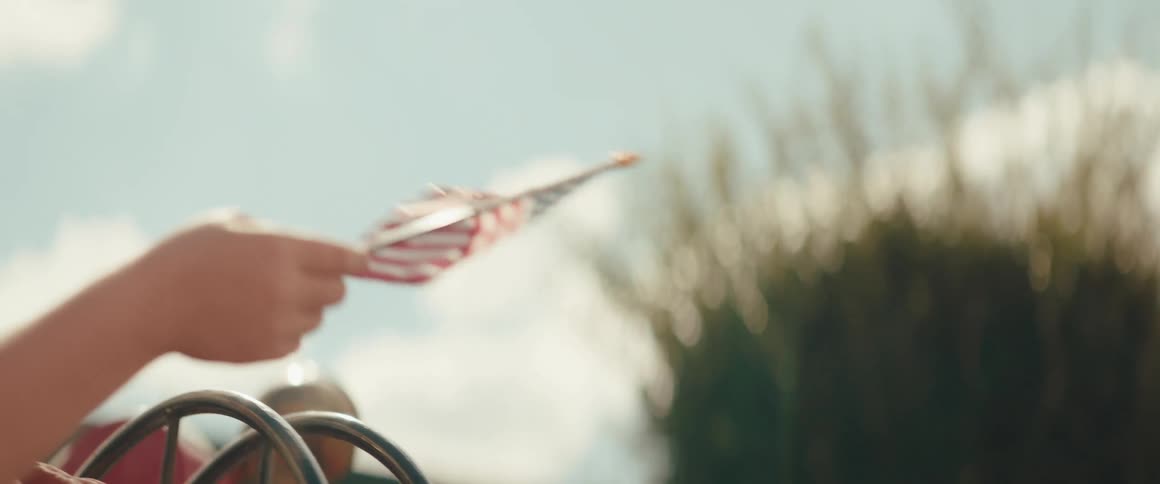 A persons hand waving a small united states flag outdoors
