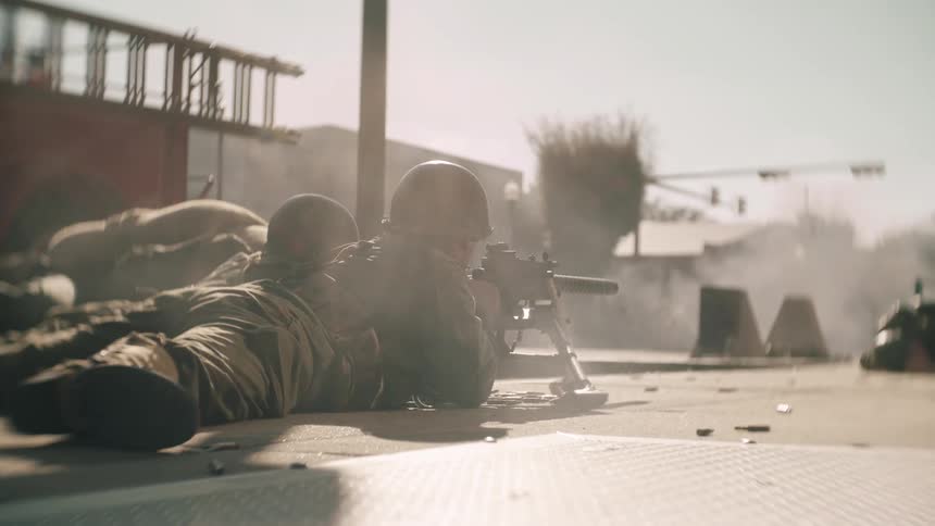 Two military officers crouched on the ground under a sunny sky wearing their uniforms and positioned behind a machine gun as they fire the gun