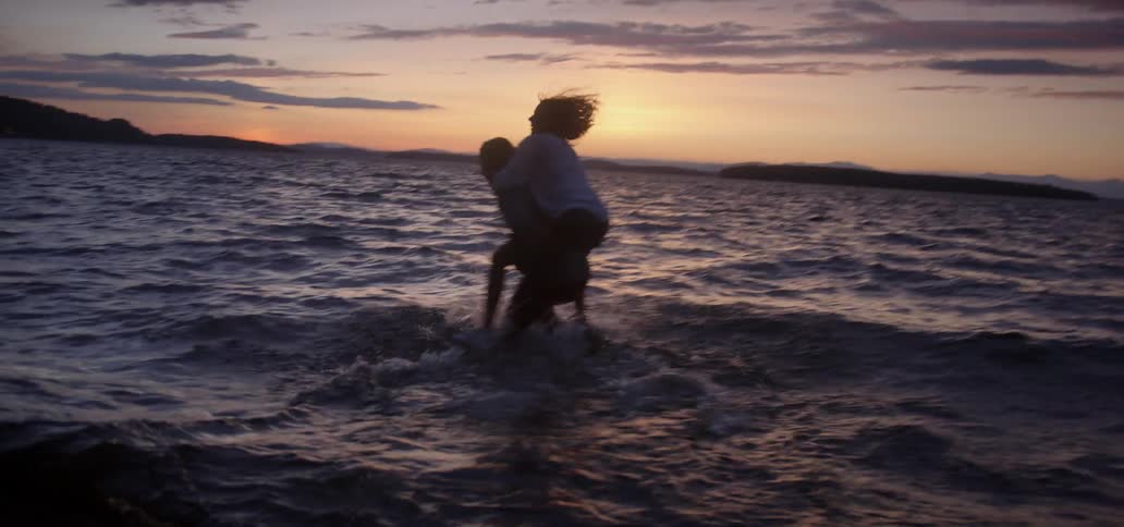 A woman rides on a man's shoulders as he runs into the ocean at sunset