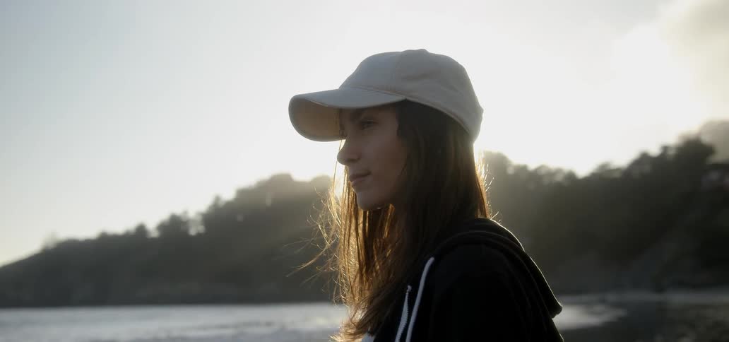 A woman stands on a shore with sunlight streaming behind her