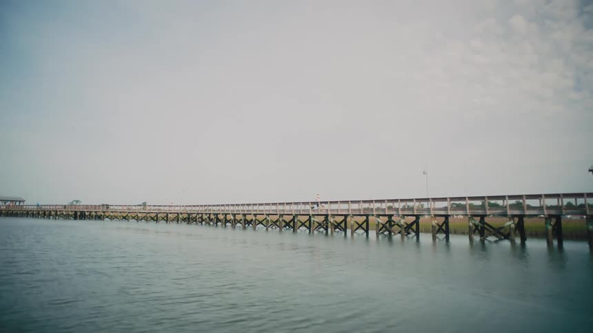 A long wooden bridge stretching across a river at midday with a person standing on a boat docked beside the bridge