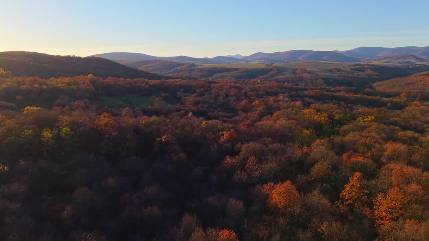 An aerial shot descends over a wide autumn colored valley and hills revealing a secluded building nestled within the forest