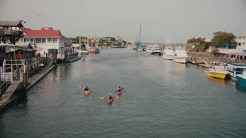 Three people in kayaks paddle down a wide waterway lined with moored white boats and colorful shops capturing an outdoor recreational scene in a lively coastal harbor town under an overcast sky