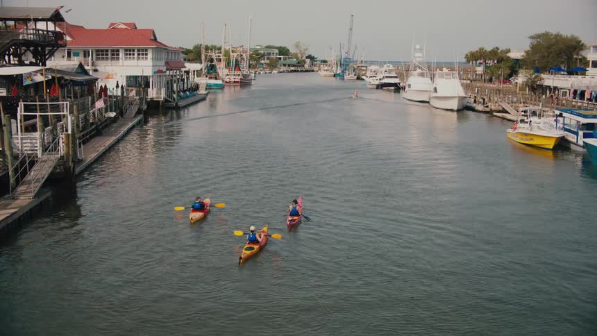 Three people kayaking through a canal at midday with boats docked along both sides of the water