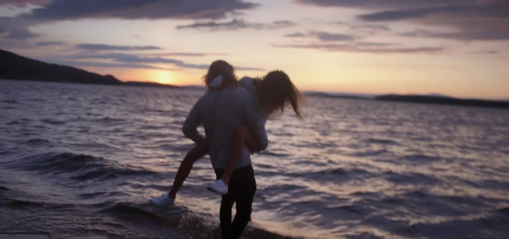 A man on the beach hugs a woman and carries her into the water at sunset