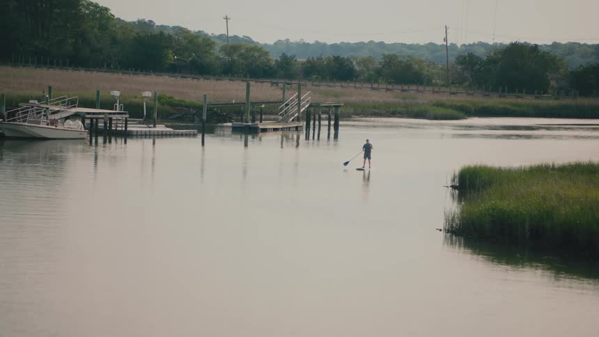 A lone paddleboarder navigates the calm waters of a coastal inlet gliding past wooden docks and swaying marsh grass under a quiet overcast sky