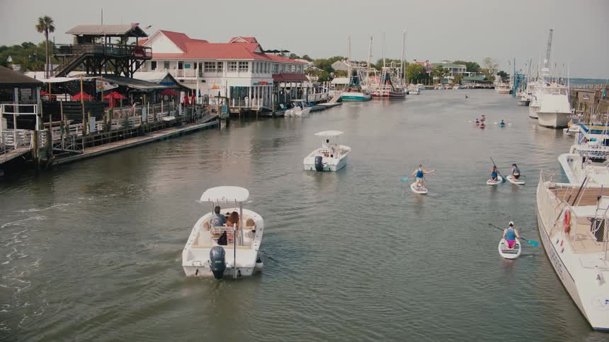 A series of boats and canoes moving through a city canal at midday with additional boats docked along the sides of the water