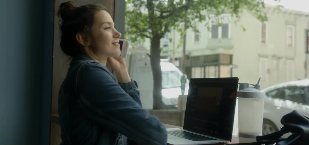A woman by a window in a cafe working on her laptop and answering her phone
