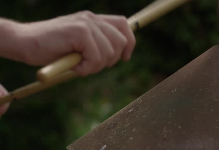 A closeup shows a persons hands holding two wooden drumsticks rapidly tapping a large flat reddishbrown rusted metal surface against a dark background