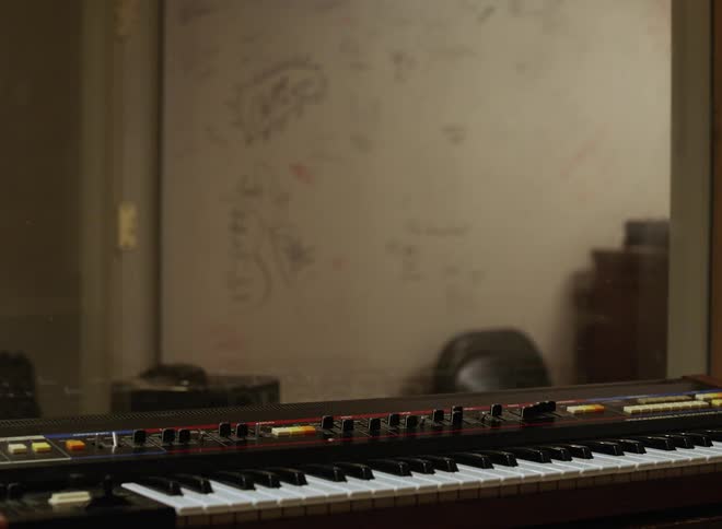 A vintage synthesizer keyboard resting in the foreground with a large reflective glass partition in the background separating a control room from a studio area and a person passing by