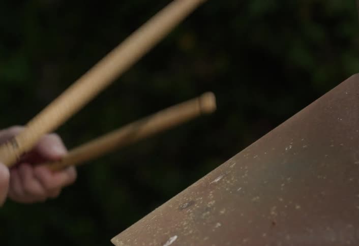 Hand holding drumsticks playing worn metal drum with blurred background