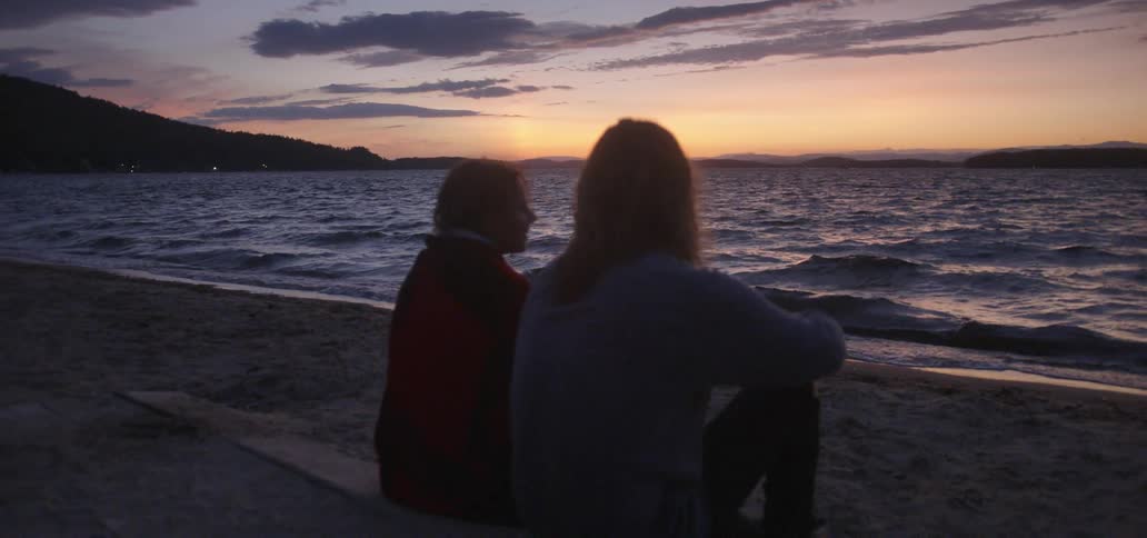 A man and woman sit on lakeshore at sunset