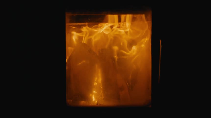 A close view shows bright orange and yellow flames intensely burning inside a dark enclosed furnace or wood stove