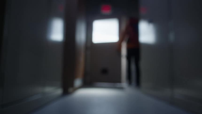 A blurred figure walks away down a dark hallway and exits through a doorway with a bright window and a red exit sign above it