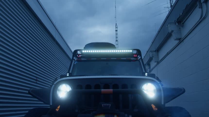 The headlights and roof light bar of a jeepstyle vehicle dramatically switch on in a narrow alley between two industrial buildings under a overcast sky