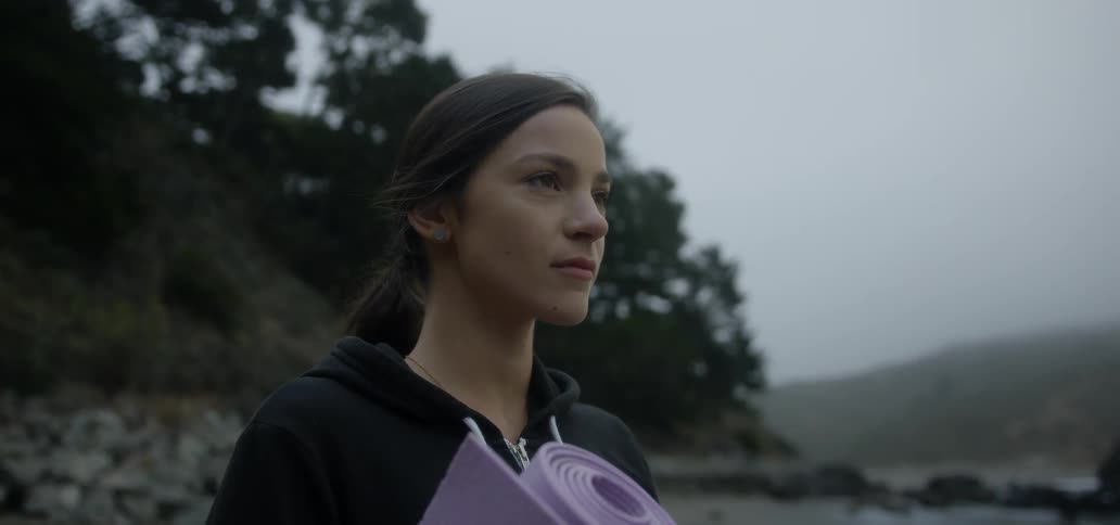 A woman holding a yoga mat outside and closes her eyes as she takes a deep breath