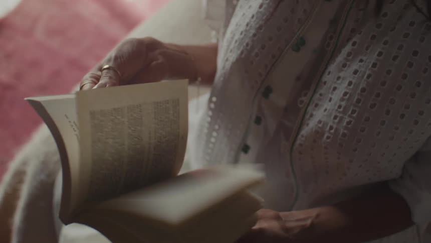 A woman sitting on her bed at midday wearing a white gown as she flips through the pages of a book
