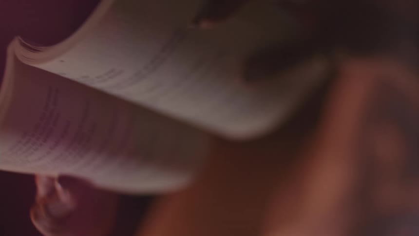 A closeup shot under warm low light shows a hand holding a thick book and lifting it revealing blurred lines of text