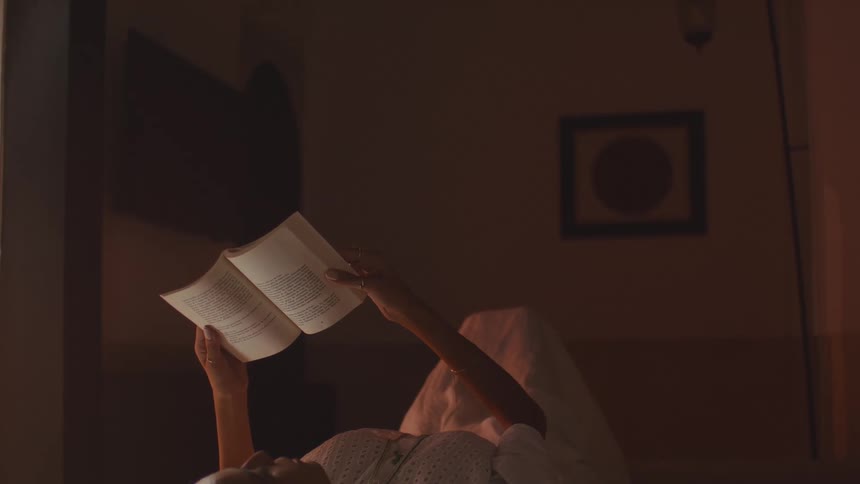 In a dimly lit quiet room a woman lies in bed holding a book overhead as its pages catch the faint warm light