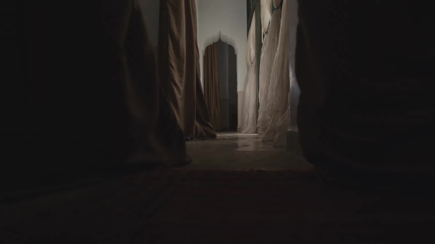 A lowangle tracking shot glides slowly down a dimly lit hallway flanked by heavy hanging drapes moving toward a bright arched doorway in the distance