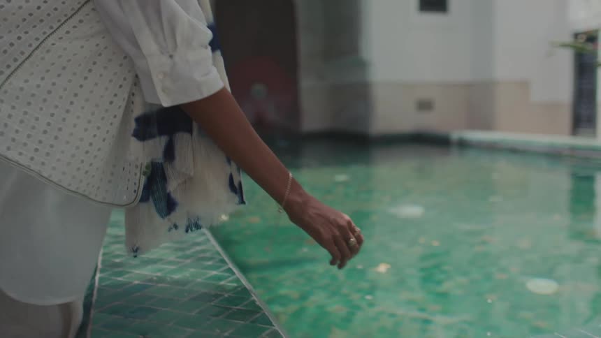 A woman in a white outfit and scarf leans down to gently touch the surface of a clear greentiled reflection pool in a bright tranquil courtyard with traditional architecture in the background
