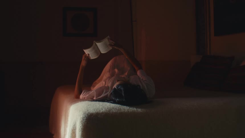 Person lying on bed holding open book above face in softly lit warm setting