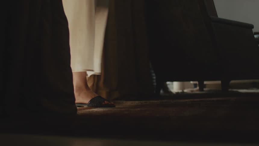 Feet wearing sandals appear beneath curtains close to the floor in an indoor setting