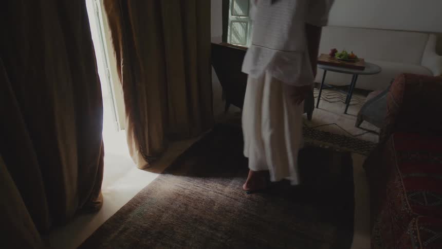 A woman wearing a white gown walking in from outdoors into a living room moving toward a couch to sit