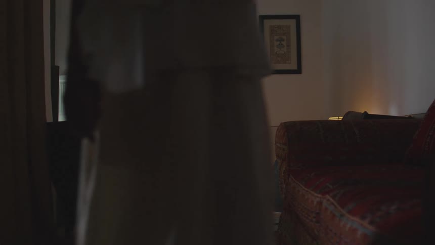A woman in a light robe quickly moves from right to left obscuring a dimly lit living room furnished with armchairs and a small table with fruit as she passes in front of a slightly open doorway