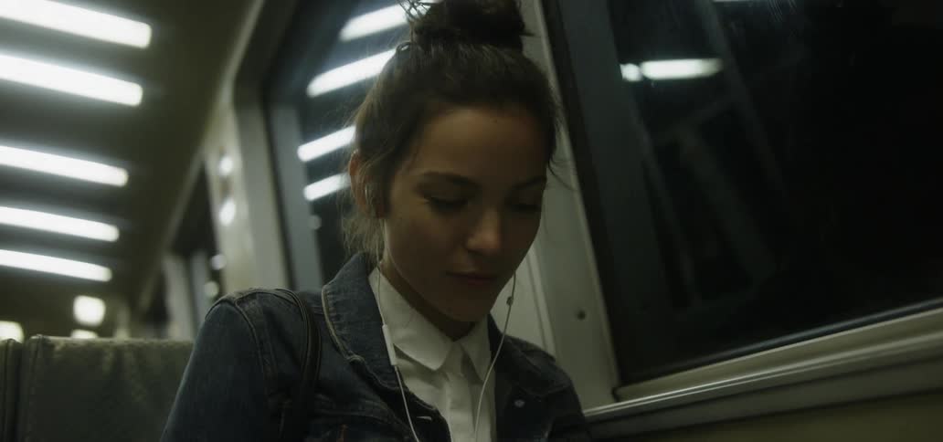 A woman on a subway train at night wearing ear buds and looking down