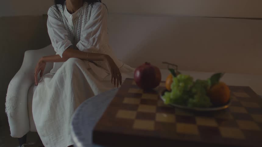 A woman in a white dress sitting in a white armchair positioned behind a round table holding a chessboard and a still life of fruit