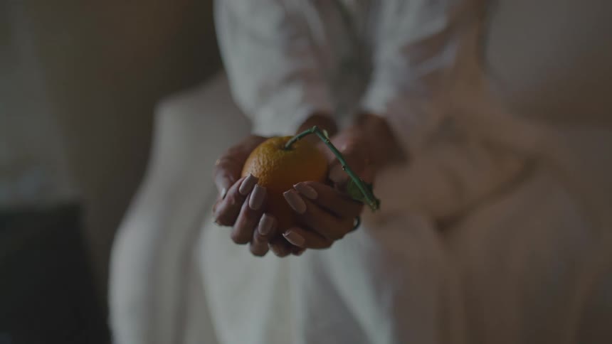 Person seated on light couch holding orange fruit with green leaves in offering gesture