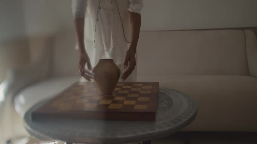 Person holds a ceramic vase above a table with a board in an indoor setting